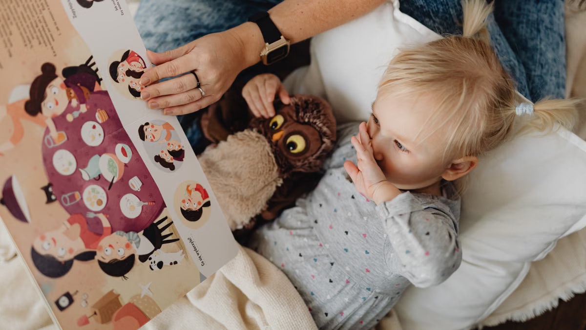 A young girl reads a storybook with her parent and a cuddly owl toy, sharing a cozy bonding moment.